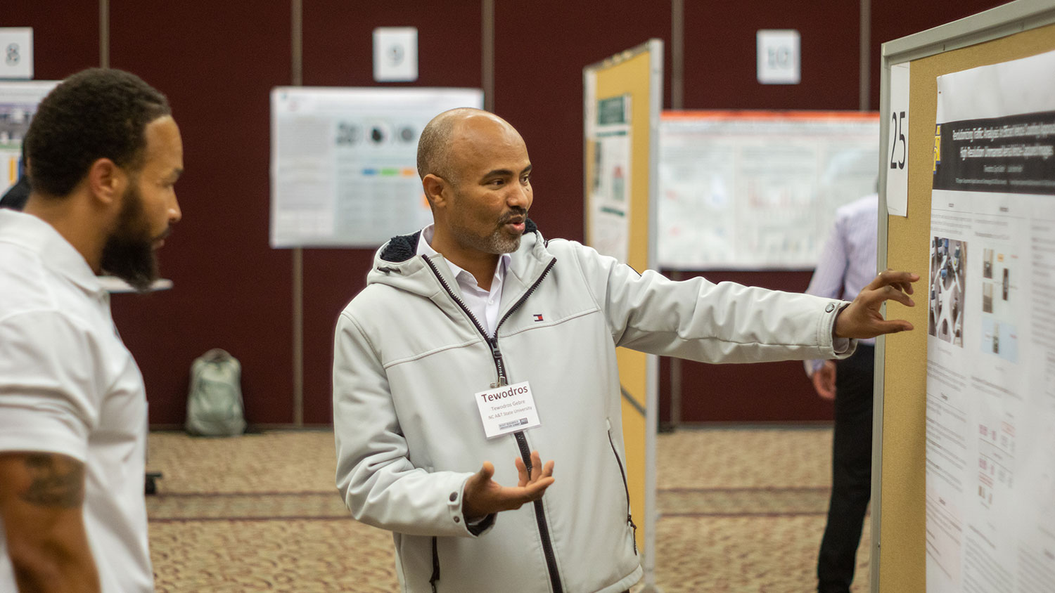 A person with medium-dark skin, a bald head, and short goatee gestures at a research poster, as a person with dark skin, short brown hair, and a brown beard watches intently